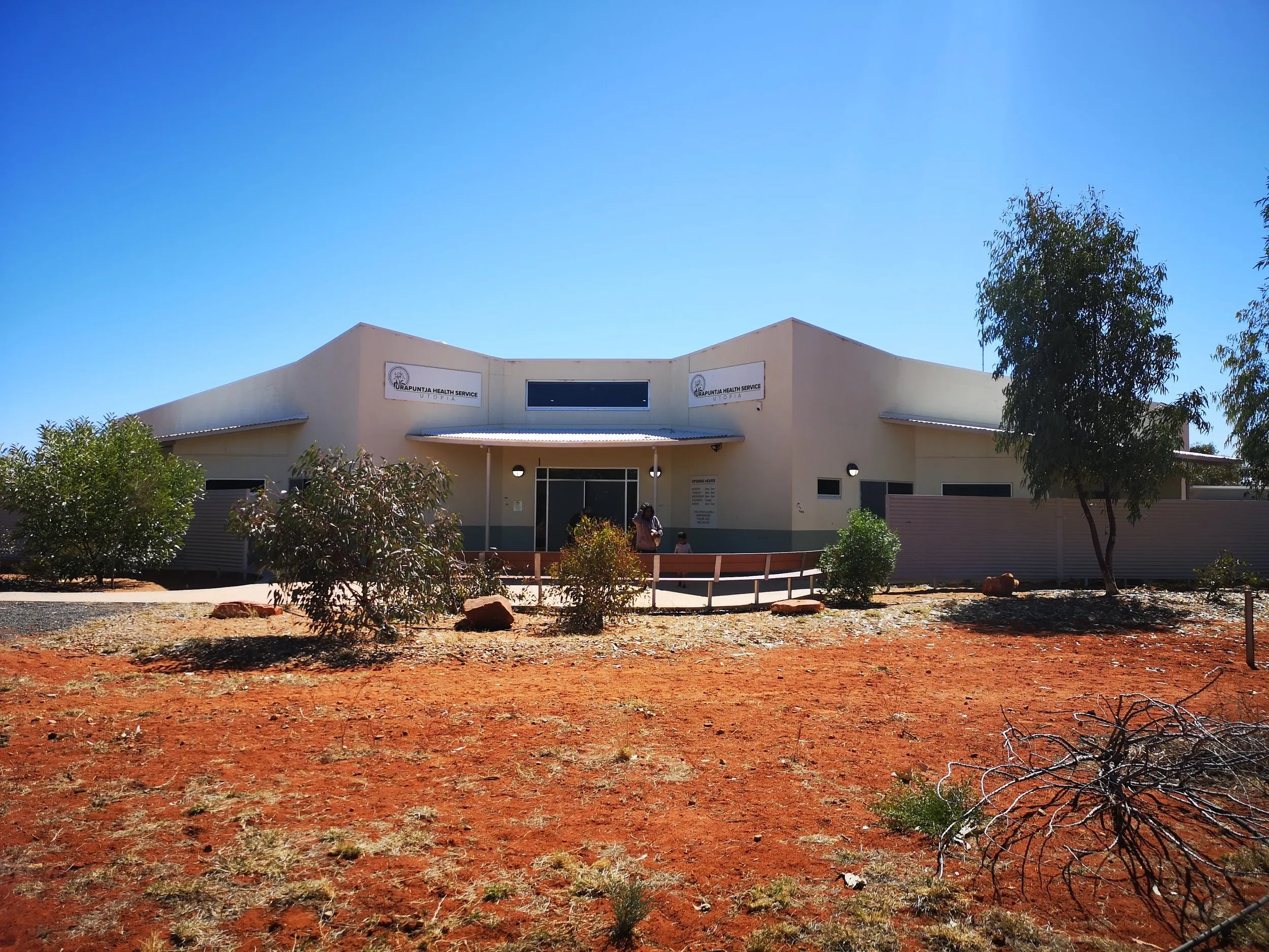 The Urapuntja Health Service clinic at Amengernterneah: a low cream building with the service's signage, under a clear blue sky on red earth