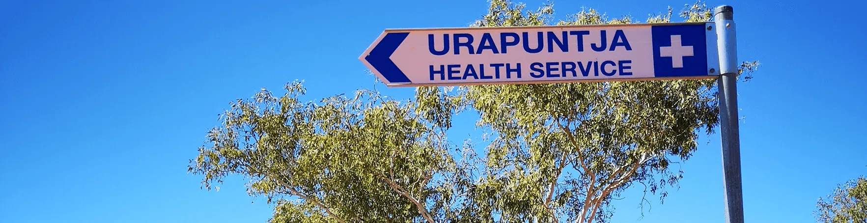 Road sign against a blue sky: Urapuntja Health Service, with an arrow pointing left