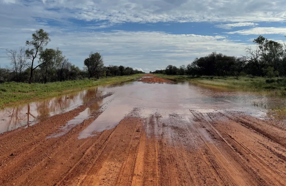The Sandover Highway after rain: a red dirt road running through grass and low gums under a wide sky, water pooled across the track